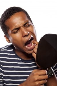 Man wearing a striped shirt touching his tooth as he looks into a handheld mirror.